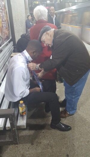 Para staruszków uczy młodego mężczyznę, jak wiązać krawat.